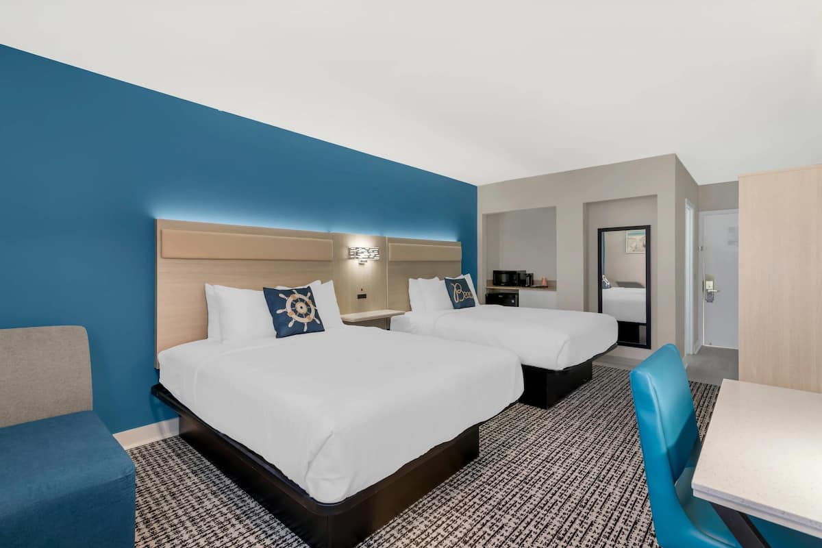 A hotel room with two beds, a desk, a chair, and a small kitchenette.