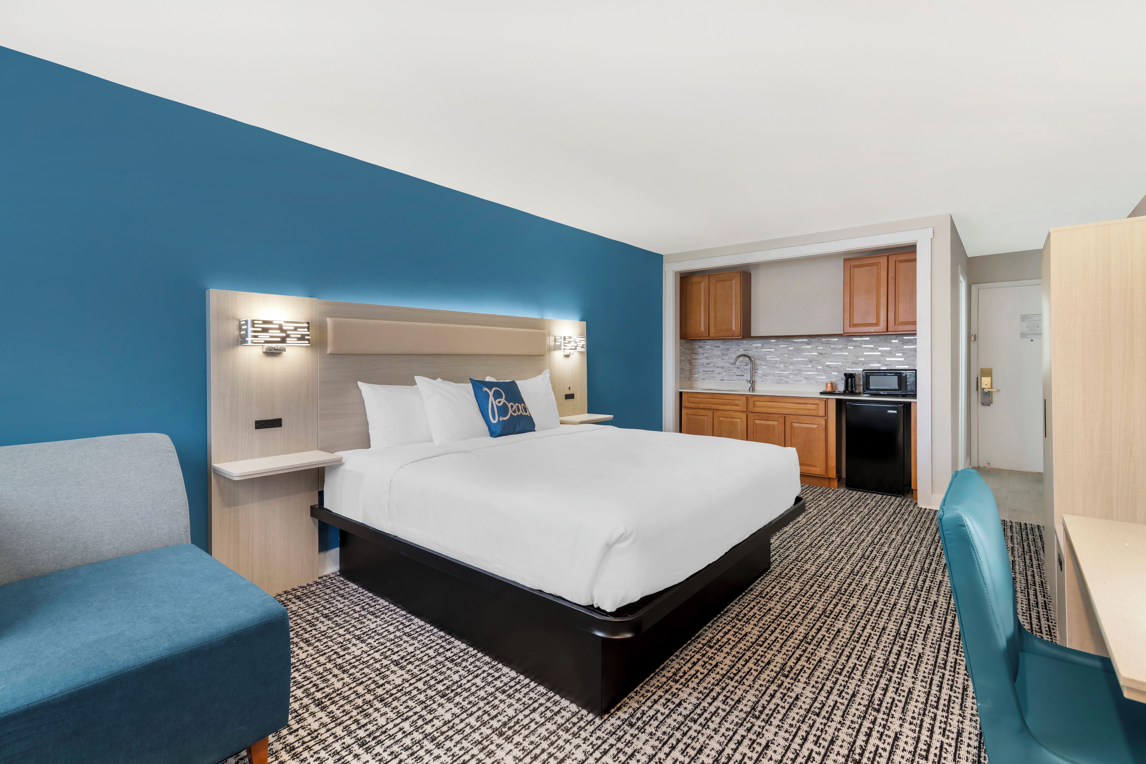 A hotel room with a bed, a desk, a chair, a kitchenette, and a bathroom.
