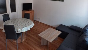 En hyggelig opholdsstue med et rundt bord, to stole, en sofa, et fjernsyn på et træskab og et billede på væggen.