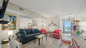 A living room with a sofa, chairs, a coffee table, and a dining table with chairs. There is a painting on the wall and a chandelier. - Oceanfront Condo, Sanabel 505: 2BR 2BA with Outdoor Hot tub (North Myrtle Beach)