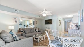 A living room with a sofa, coffee table, and dining table with chairs. There is a ceiling fan, a flat-screen TV, and artwork on the walls. - Sanabel 506, Affordable Oceanfront 2BR 2BA condo (North Myrtle Beach)