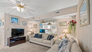 A living room with a beige sofa, a TV on a wooden stand, a ceiling fan, and a kitchen in the background. - Sanabel 609: Oceanfront 2BR/2BA- Indoor Pool (North Myrtle Beach)