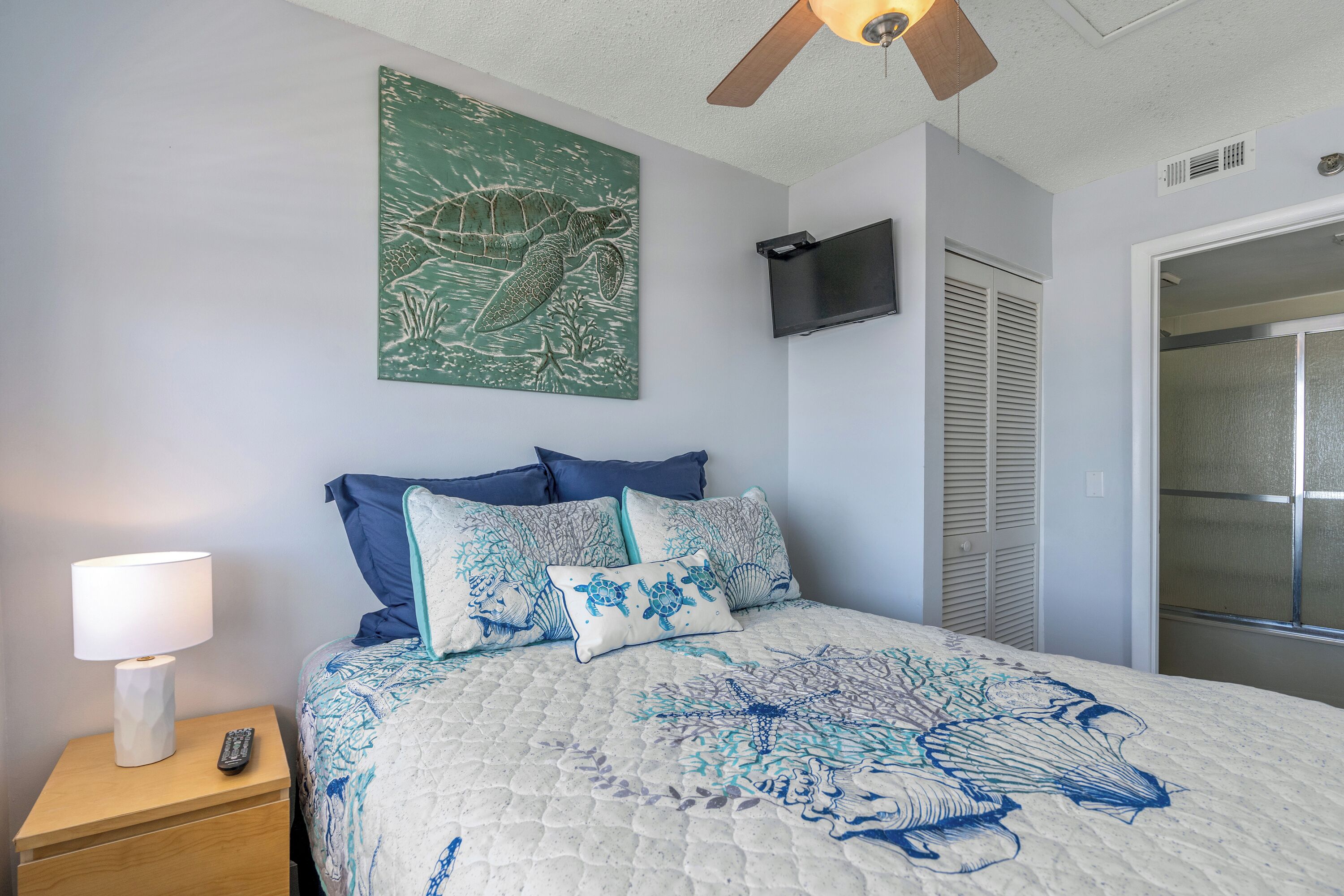 Une chambre à coucher comprenant un lit, une table de chevet, un téléviseur fixé au mur et un ventilateur de plafond.