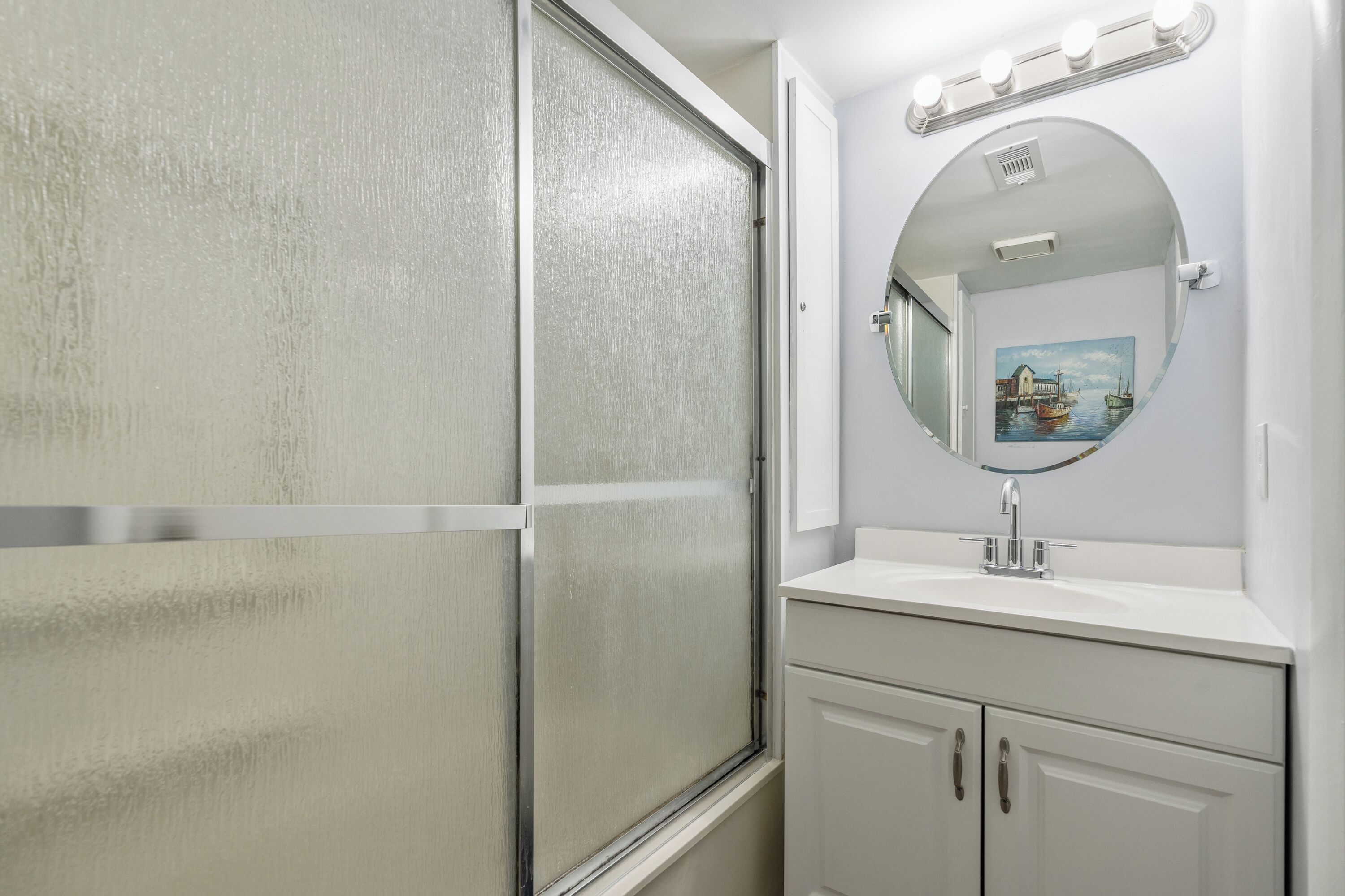 A bathroom with a frosted glass shower door, a round mirror with lights, and a sink with cabinets.
