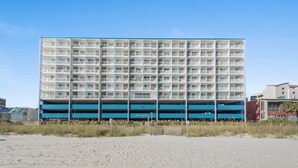 A multi-story hotel with a blue and white facade, located on a sandy beach. - Sanabel 215, Extraordinary Views from Extra Large Oceanfront Balcony (North Myrtle Beach)