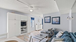 A modern living room with a sofa, a dining table, and a TV. There is a ceiling fan and a starfish decoration on the wall. - Sanabel 215, Extraordinary Views from Extra Large Oceanfront Balcony (North Myrtle Beach)