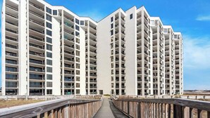 Een modern meerlaags gebouw met balkons, een houten looppad ernaartoe en een heldere lucht.