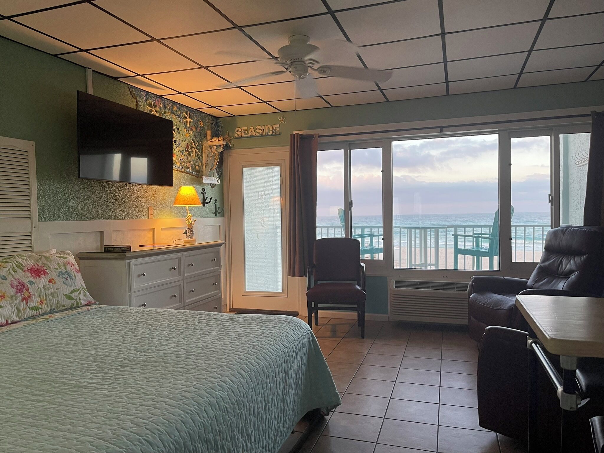 A bedroom with a bed, a desk, a chair, a TV, a lamp, and a view of the ocean.