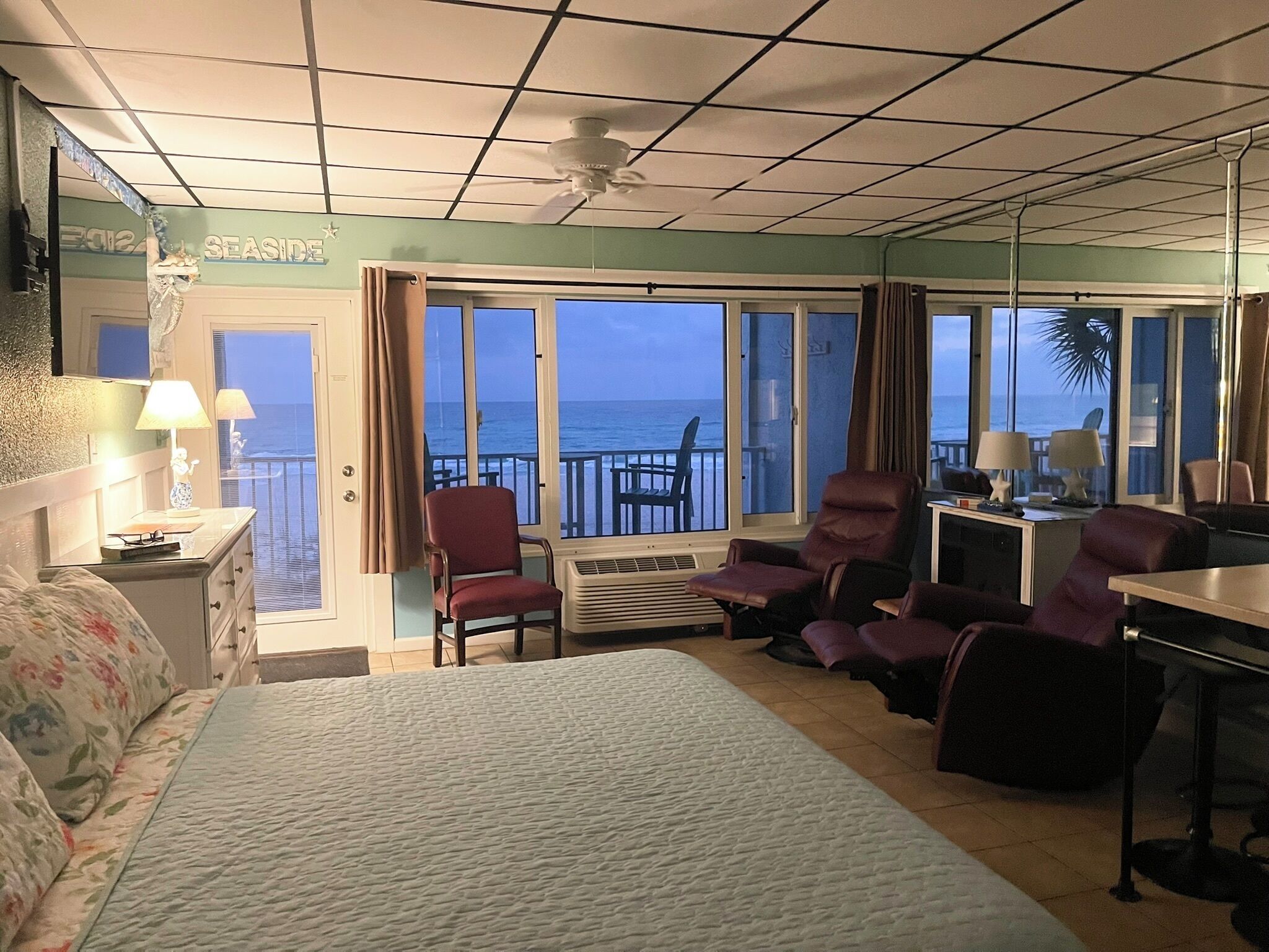 A hotel room with a bed, two armchairs, a desk, a computer, and large windows with a view of the ocean.