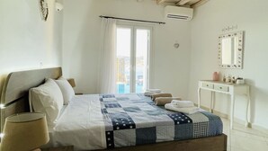 Un dormitorio con techo de madera, una cama con colcha, una mesita de noche con lámpara, un pequeño aparador blanco y una ventana con cortinas.