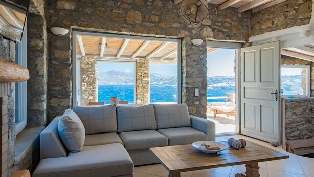 Habitación con paredes de piedra y un amplio ventanal con vistas al mar y a las montañas.