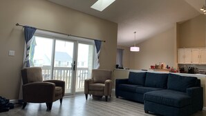 Un vaste salon doté d’une grande porte-fenêtre coulissante donnant sur un balcon, un coin cuisine avec des meubles blancs et un canapé bleu.