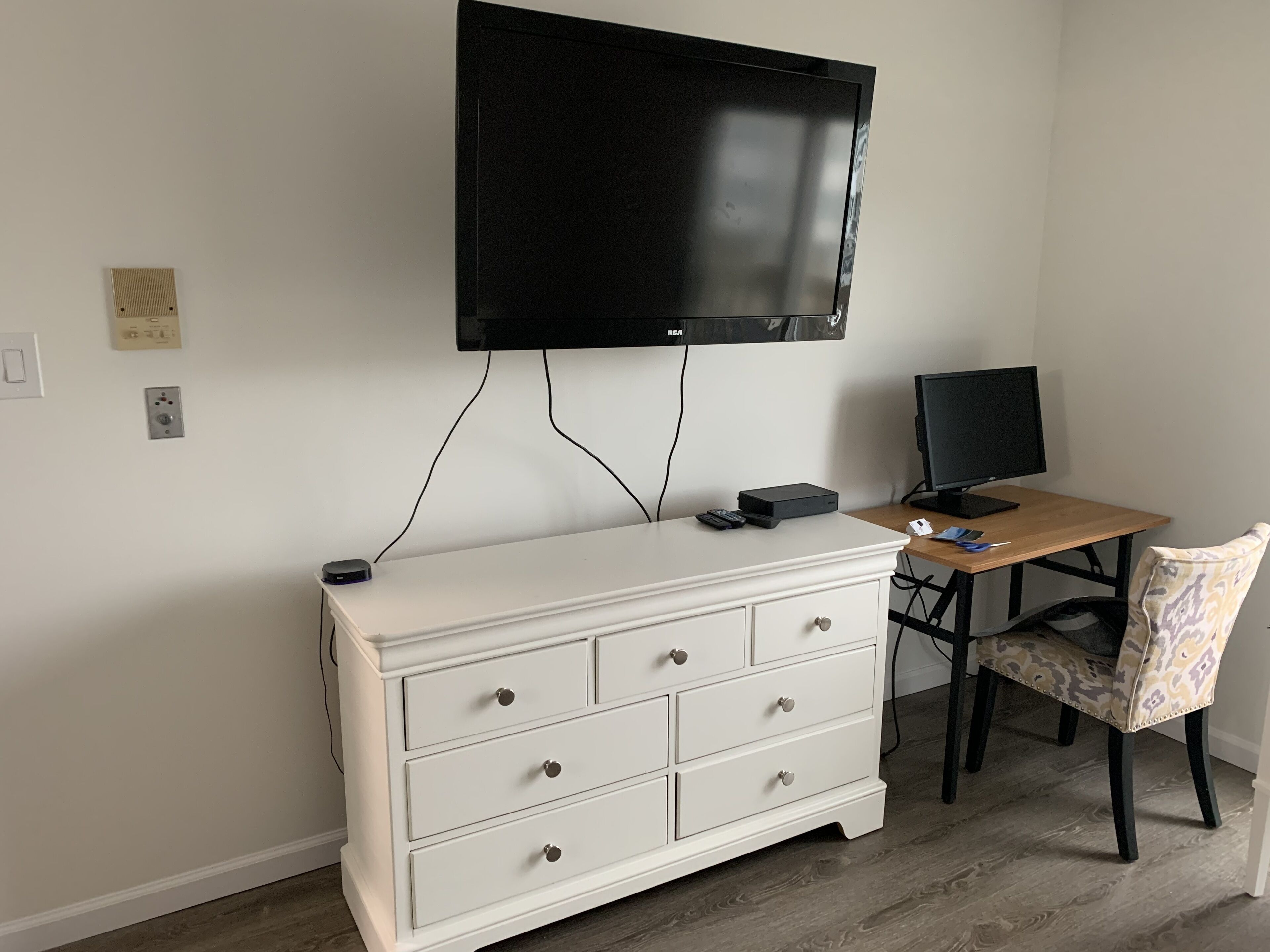 A room with a white dresser, a desk with a computer, and a TV mounted on the wall.