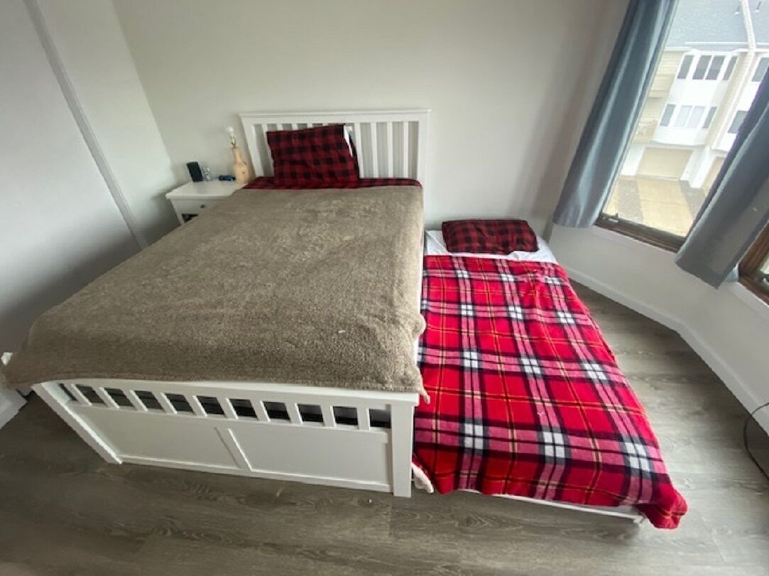Un lit simple avec une tête de lit et un pied de lit blancs, une couverture à carreaux rouges et noirs, et un plaid beige.