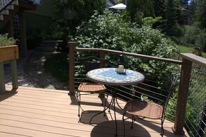 Une terrasse en bois avec une table ronde et deux chaises en osier, entourée de verdure.