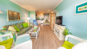 A living room with a sofa, chairs, a coffee table, a TV, and a kitchen in the background. - Sea Dreams: Affordable Family Friendly: Ocean Views • Pool • Linens & Amenities (Atlantic Beach)