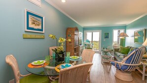 A dining area with a glass table, wicker chairs, and a bookshelf. - Sea Dreams: Affordable Family Friendly: Ocean Views • Pool • Linens & Amenities (Atlantic Beach)
