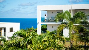 A modern building with a balcony overlooking the sea, surrounded by lush greenery.