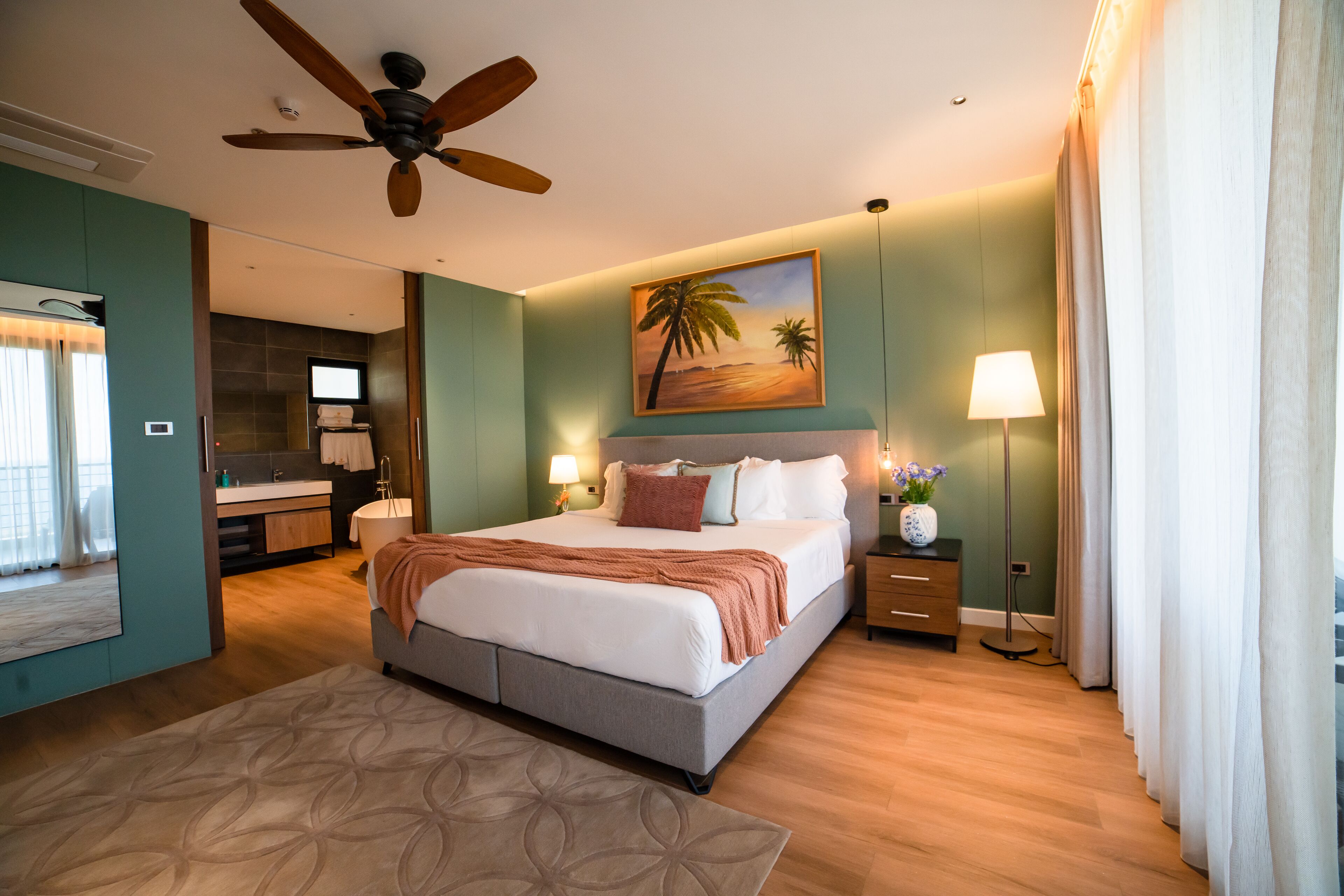 Una habitación de hotel moderna con una cama grande, un ventilador de techo, un cuadro de una playa, un baño con un espejo amplio y una mesita de noche con una lámpara.