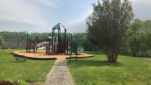 Un terrain de jeux équipé d’un toboggan, de structures d’escalade et d’un espace abrité, le tout entouré de pelouse et d’arbres.