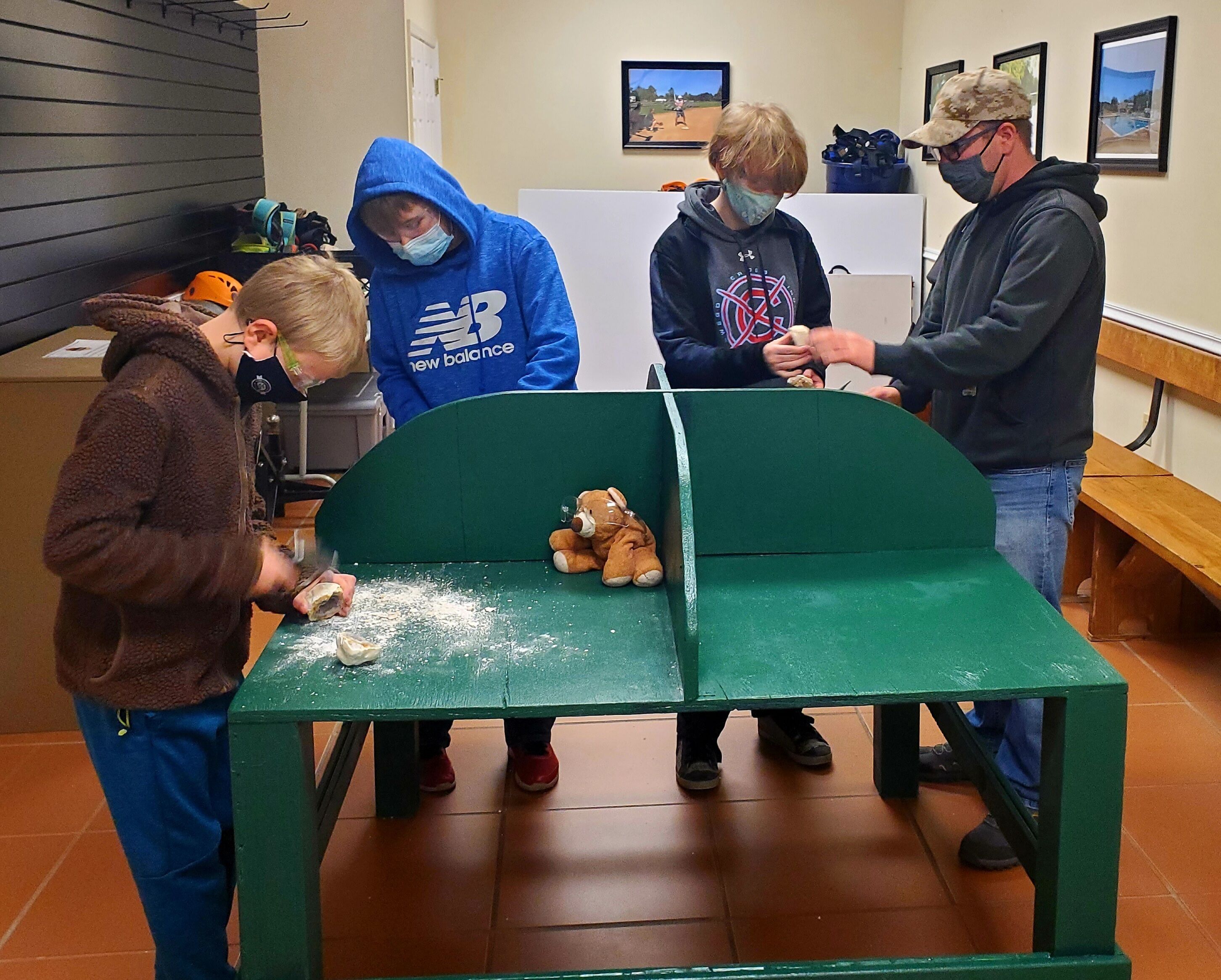 A group of people around a green bench with a teddy bear on it.
