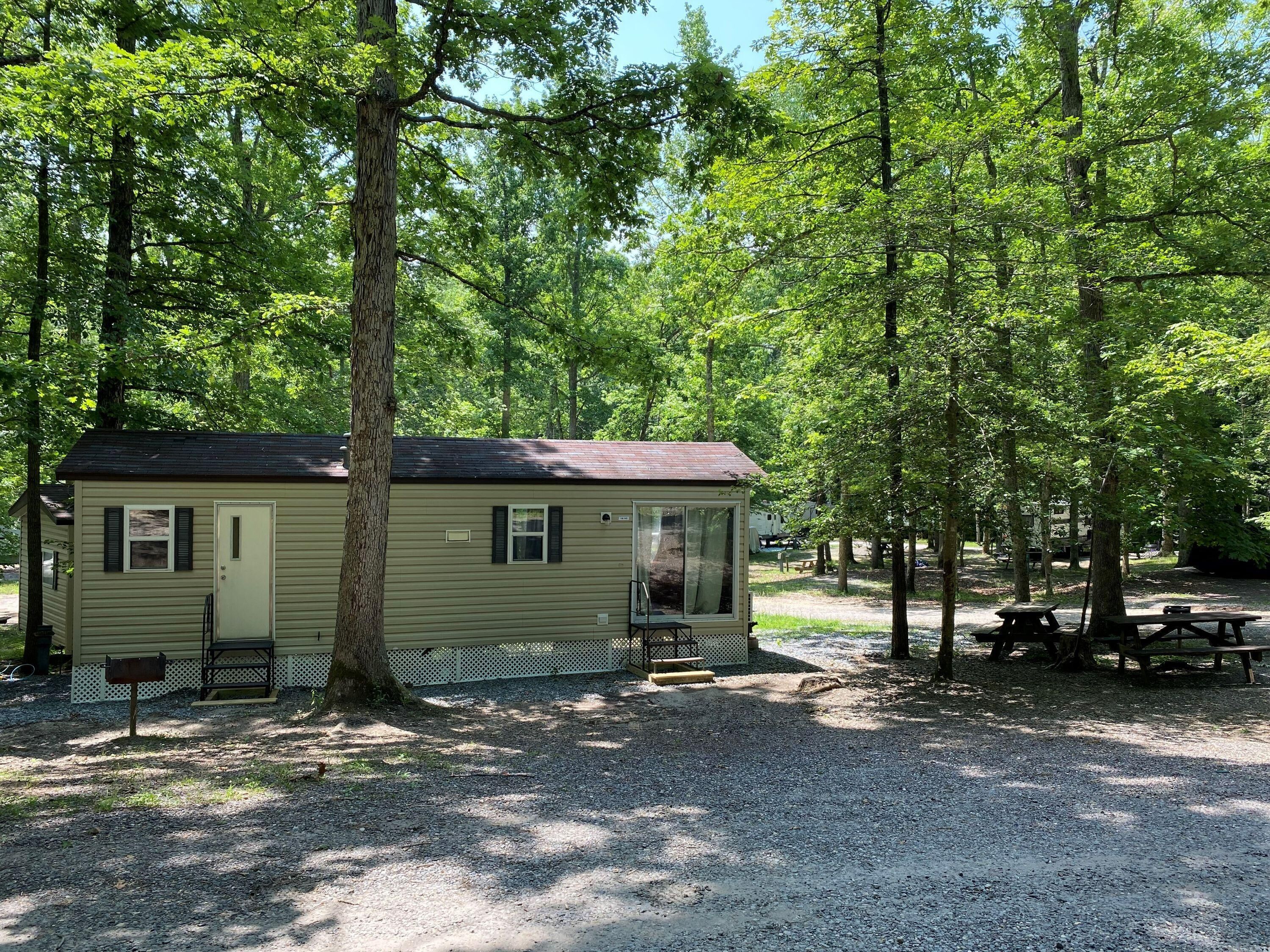 A small, single-story cabin with a porch, surrounded by trees in a campground setting.