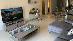 A modern living room with a flat-screen TV, a glass coffee table, and a grey sofa. There is a desk with a chair and a dining area with a glass table and chairs. - Amazing penthouse close to the beach (Cabo San Lucas)