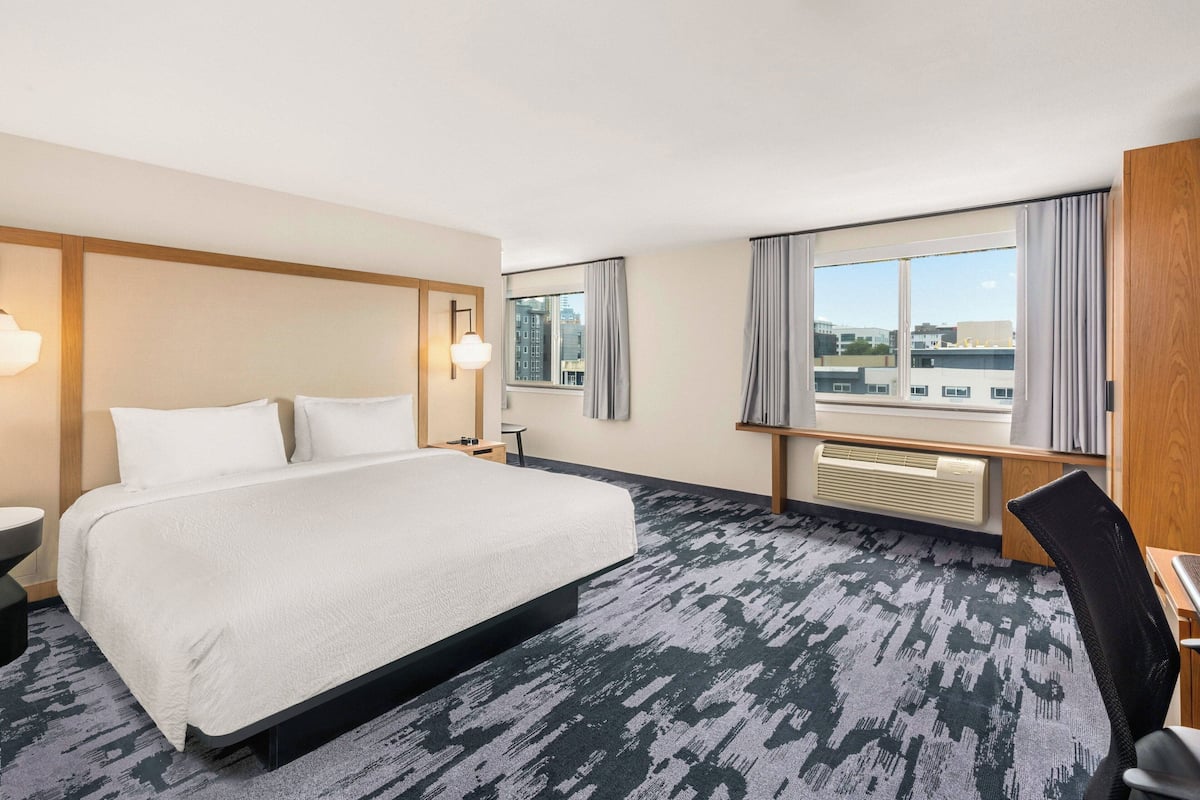 A hotel room with a large bed, a desk, a chair, a window with curtains, and a view of buildings.
