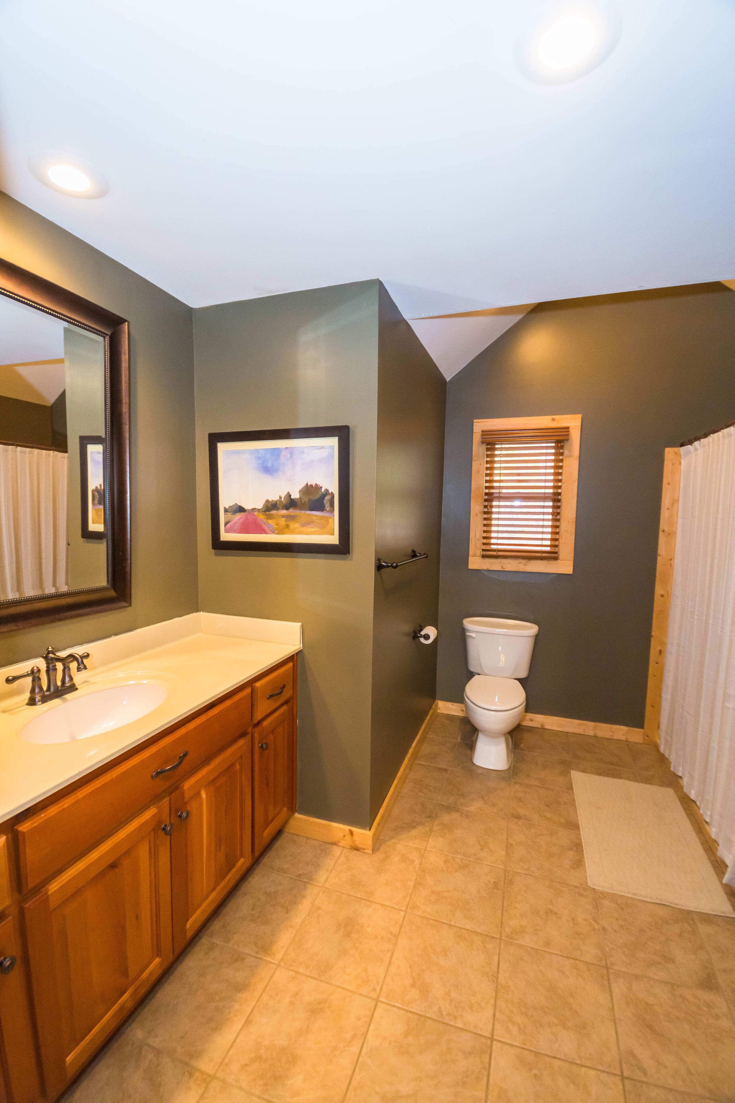 Une salle de bain avec deux lavabos, des toilettes, un tableau encadré au mur et une fenêtre avec des stores.