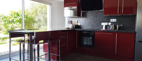 A modern kitchen with red cabinets, a black stove, and a dishwasher. There is a bar stool and a window with a view of greenery outside.