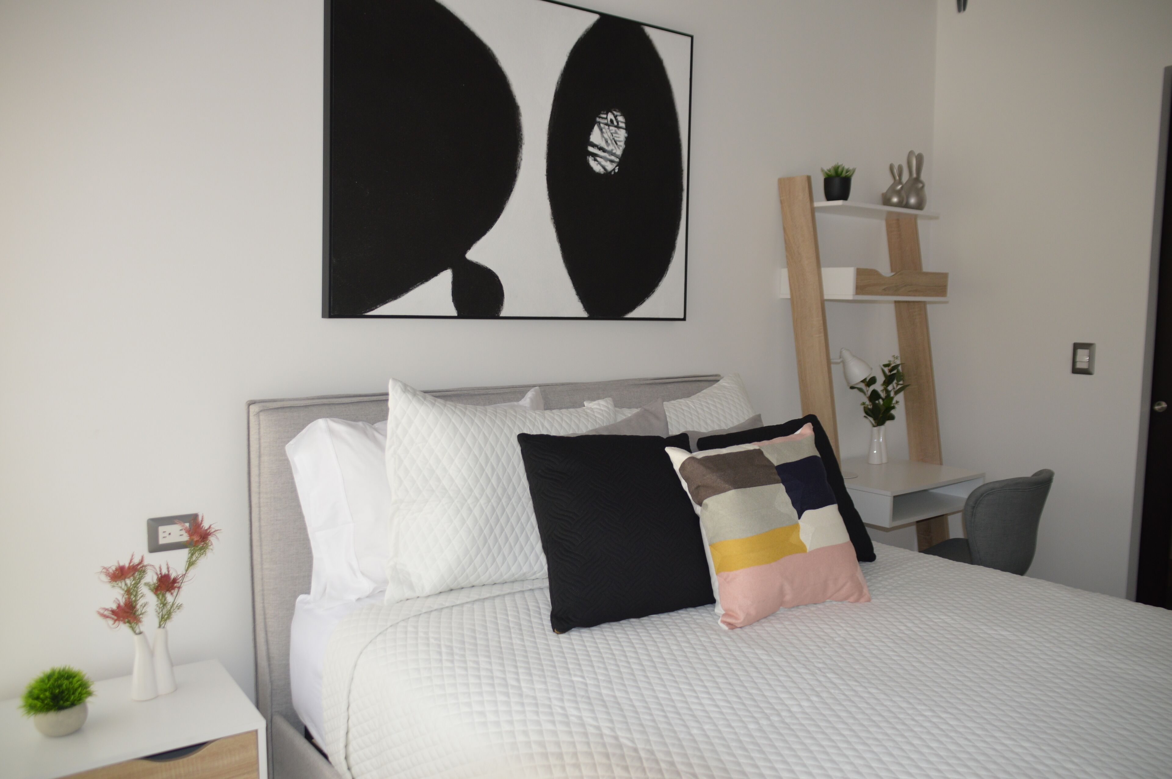 Un dormitorio ordenado con cama, almohadas, una mesita de noche con un florero y una obra de arte en la pared.