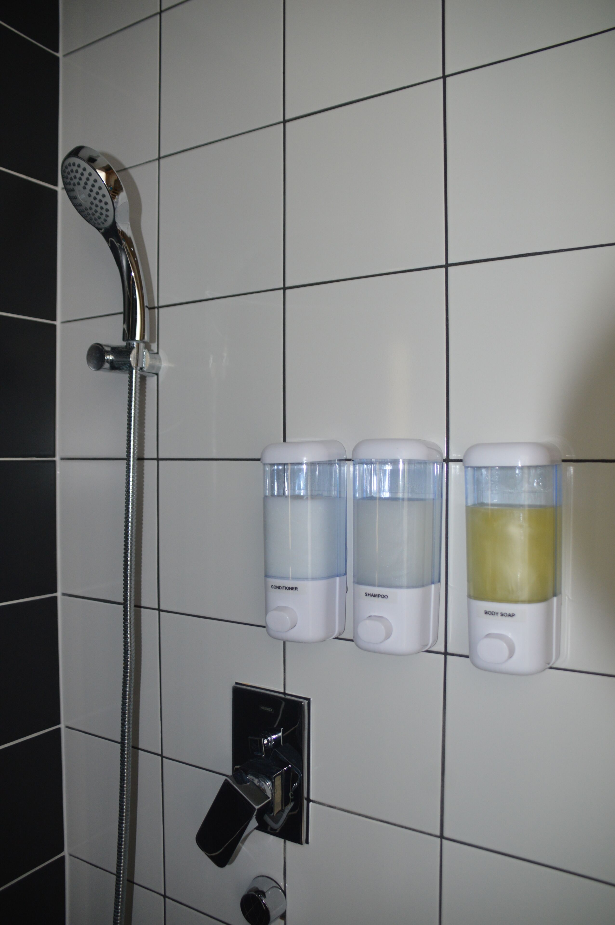 A bathroom with a shower, three dispensers for conditioner, shampoo, and body soap, and a control panel.