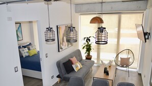 Una sala moderna con un sofá, una mesita de centro y un comedor. Hay una cama con colchón y almohadas azules, una planta en maceta y una silla.