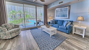 A living room with a blue sofa, a patterned armchair, a coffee table, and a view of a pool and buildings through large sliding glass doors.