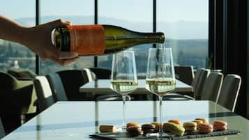 A hand pouring white wine into two glasses on a table with macarons, in a room with a view of mountains through large windows.