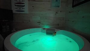 A dimly lit room with a circular hot tub in the center, surrounded by wooden walls and a small shelf with items. - The Homestead, in Bedeque, waterview beach house and hot tub cabin (Bedeque)