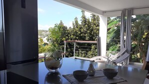 A modern kitchen with a refrigerator, a dining table with a glass bowl and two bowls, and a view of lush greenery and a red-roofed structure outside. - Magnificent and luxurious T2 bis (Les Trois Ilets)