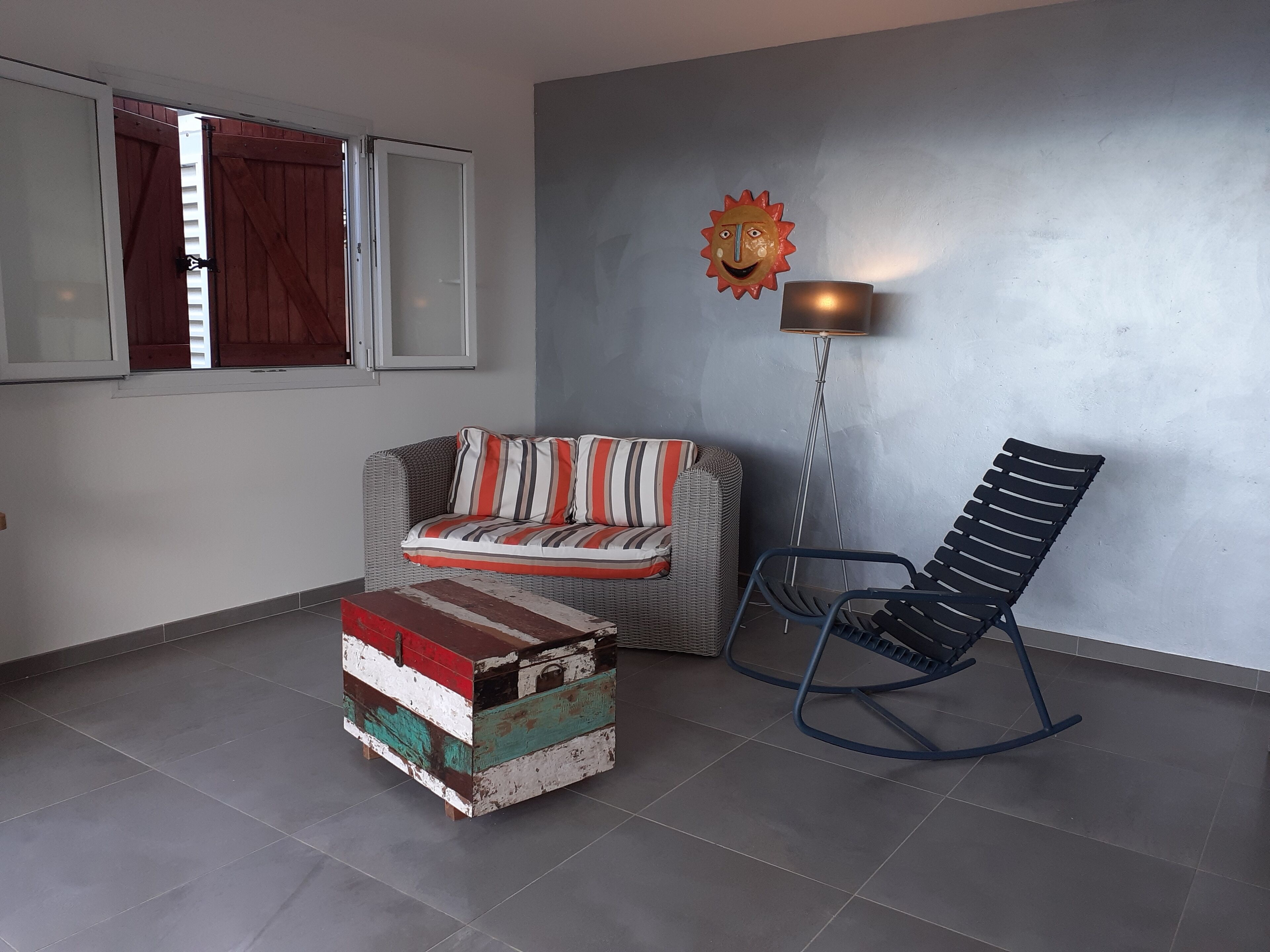 Una habitación con sofá, sillón mecedora, pequeño cofre de madera y lámpara de pie.