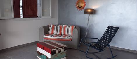 Una habitación con sofá, sillón mecedora, pequeño cofre de madera y lámpara de pie.