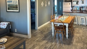 Una cocina moderna con armarios blancos, una zona de comedor con mesa y sillas de madera, y una pared gris con elementos decorativos.
