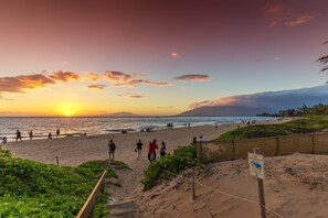 A beach with people walking and sitting, a sunset in the background, and a sign indicating dune restoration.