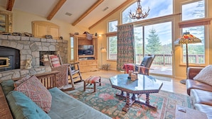 A spacious living room with a stone fireplace, wooden beams, a large window with a view of trees, and a cozy seating area. - Woodland Cabin in Lake Arrowhead: W/Two decks (Lake Arrowhead)