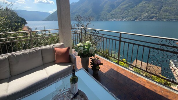 A balcony with a sofa, a table with a bottle and glasses, and a view of a lake and mountains. - Amazing Lake Views from Large Terrace.  Swimming and Boat Dock on Property (NESSO)