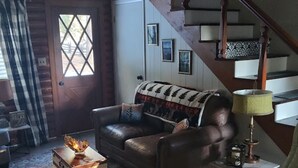 Un salon chaleureux avec un canapé en cuir marron, un tapis à motifs, une table basse en bois et un fauteuil à bascule. On aperçoit, au fond, un escalier avec des rampes en bois et une lampe.