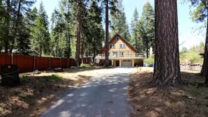 Une maison en bois à deux étages, dotée d’un porche couvert, entourée de grands arbres et bordée d’une allée en terre.