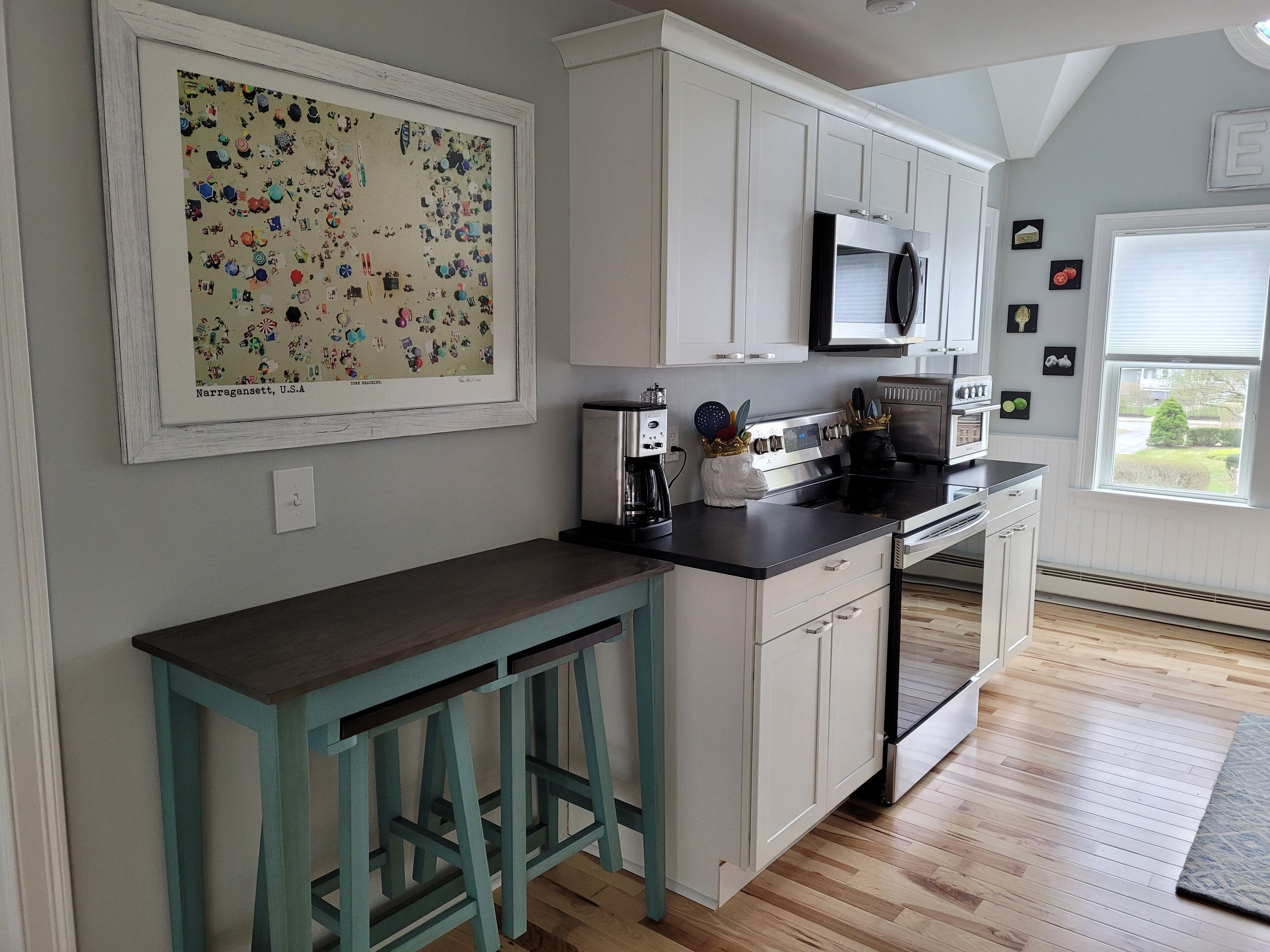A kitchen with white cabinets, a black countertop, a microwave, and a coffee maker. There is a framed picture on the wall and a window with blinds.
