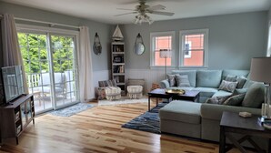 A living room with a grey sofa, wooden flooring, a ceiling fan, and a view through a sliding glass door. - Blue Moon Carriage House (Narragansett)