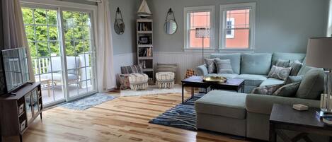 A living room with a grey sofa, wooden flooring, a ceiling fan, and a view through a sliding glass door.