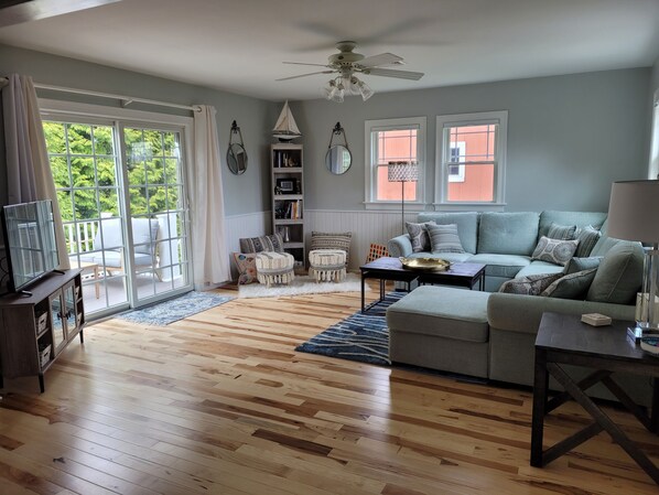 A living room with a grey sofa, wooden flooring, a ceiling fan, and a view through a sliding glass door.