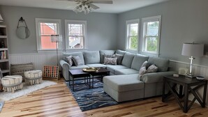 A living room with a grey sectional sofa, wooden flooring, a ceiling fan, and large windows. - Blue Moon Carriage House (Narragansett)